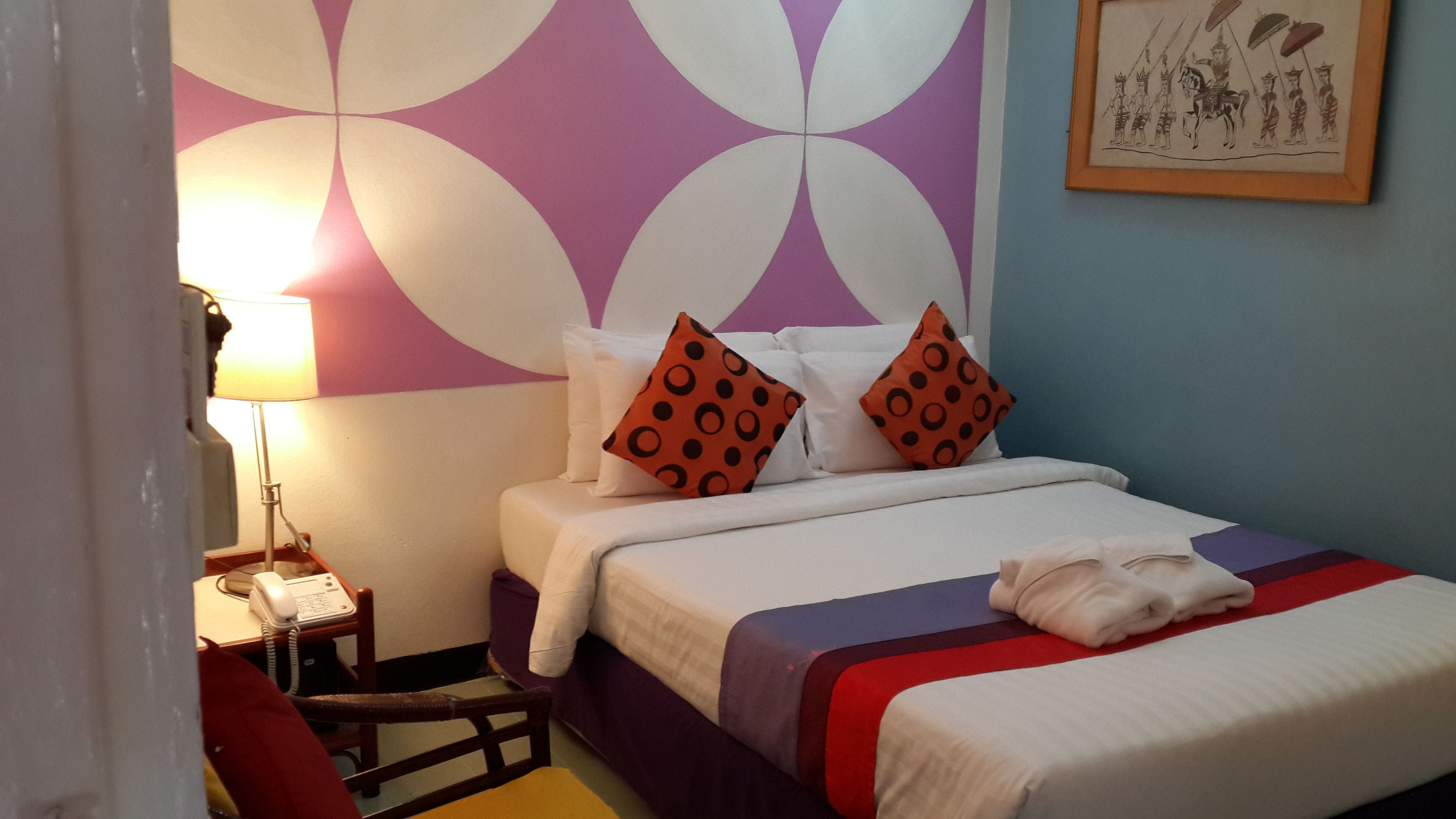 A hotel room with a bed, pillows, a nightstand, a lamp, and a wall with a geometric pattern.