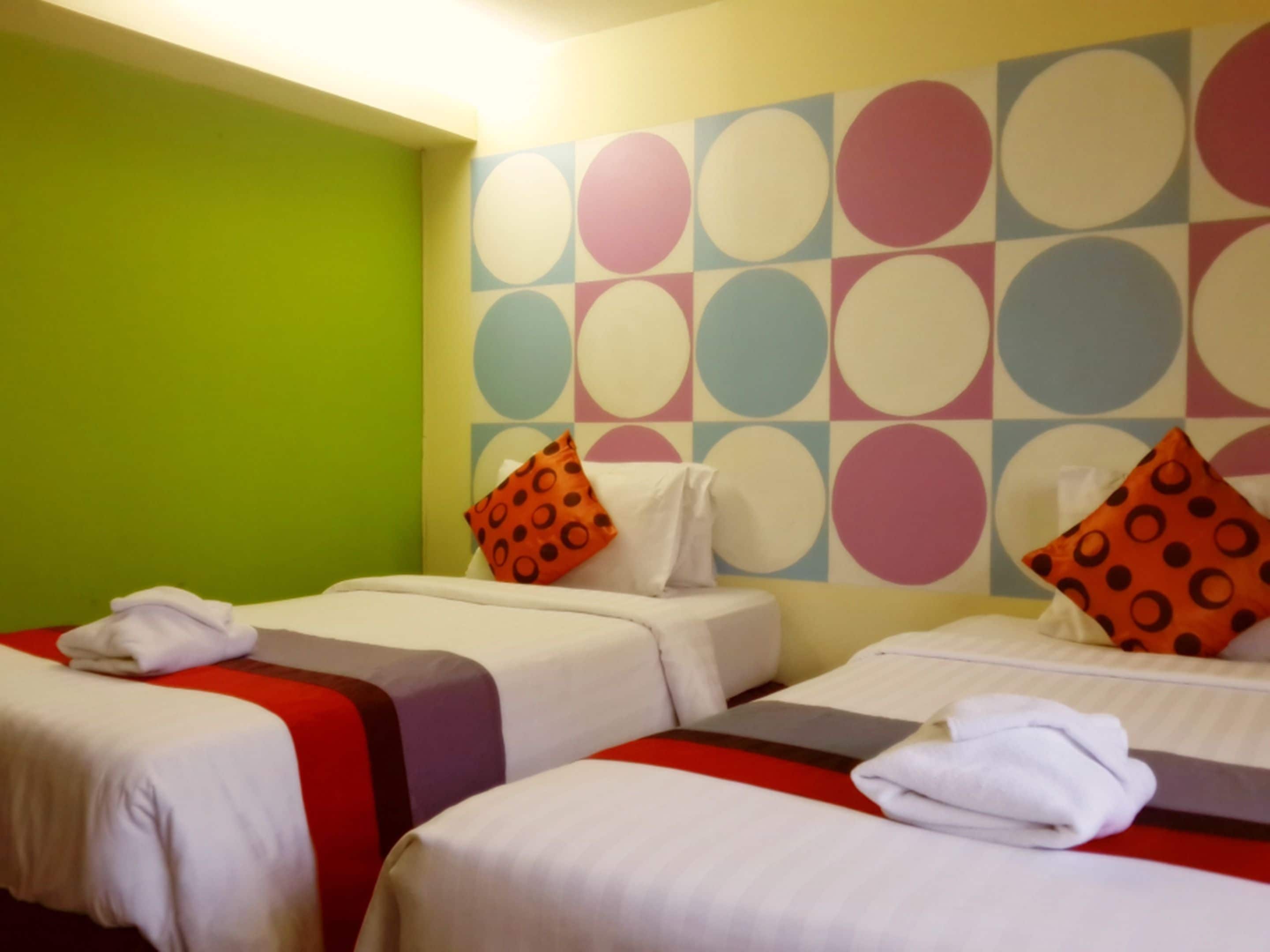 A hotel room with two beds, each with a white bedspread and a red and gray striped blanket. The walls are decorated with a colorful circular pattern.