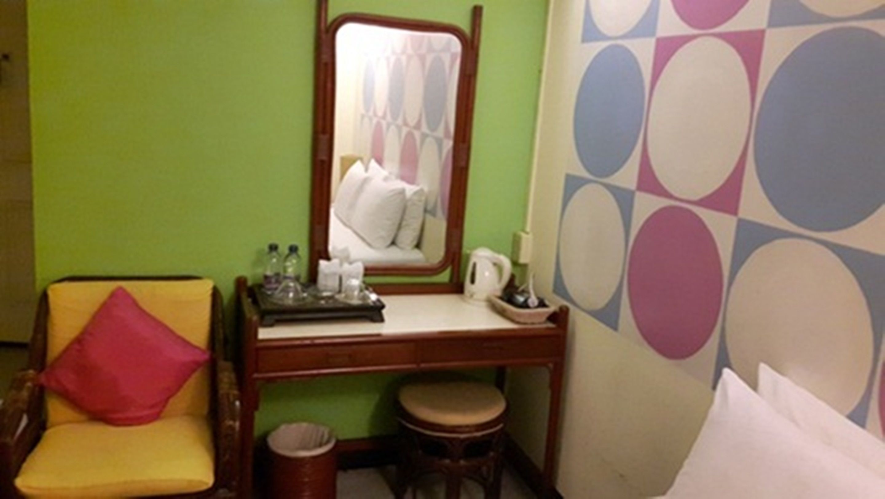 A room with a green wall, a wooden desk with a mirror, a yellow chair with a red pillow, a small stool, and a patterned wall.