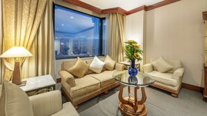 Habitación de hotel con sofá, una mesita de centro de vidrio, lámpara y vista panorámica de la ciudad a través de un ventanal.