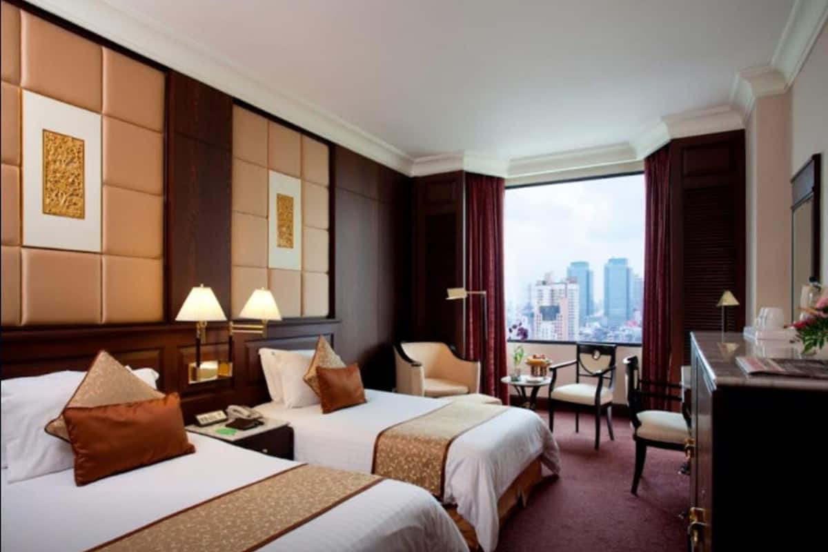A hotel room with two beds, a desk, a chair, and a view of the cityscape.