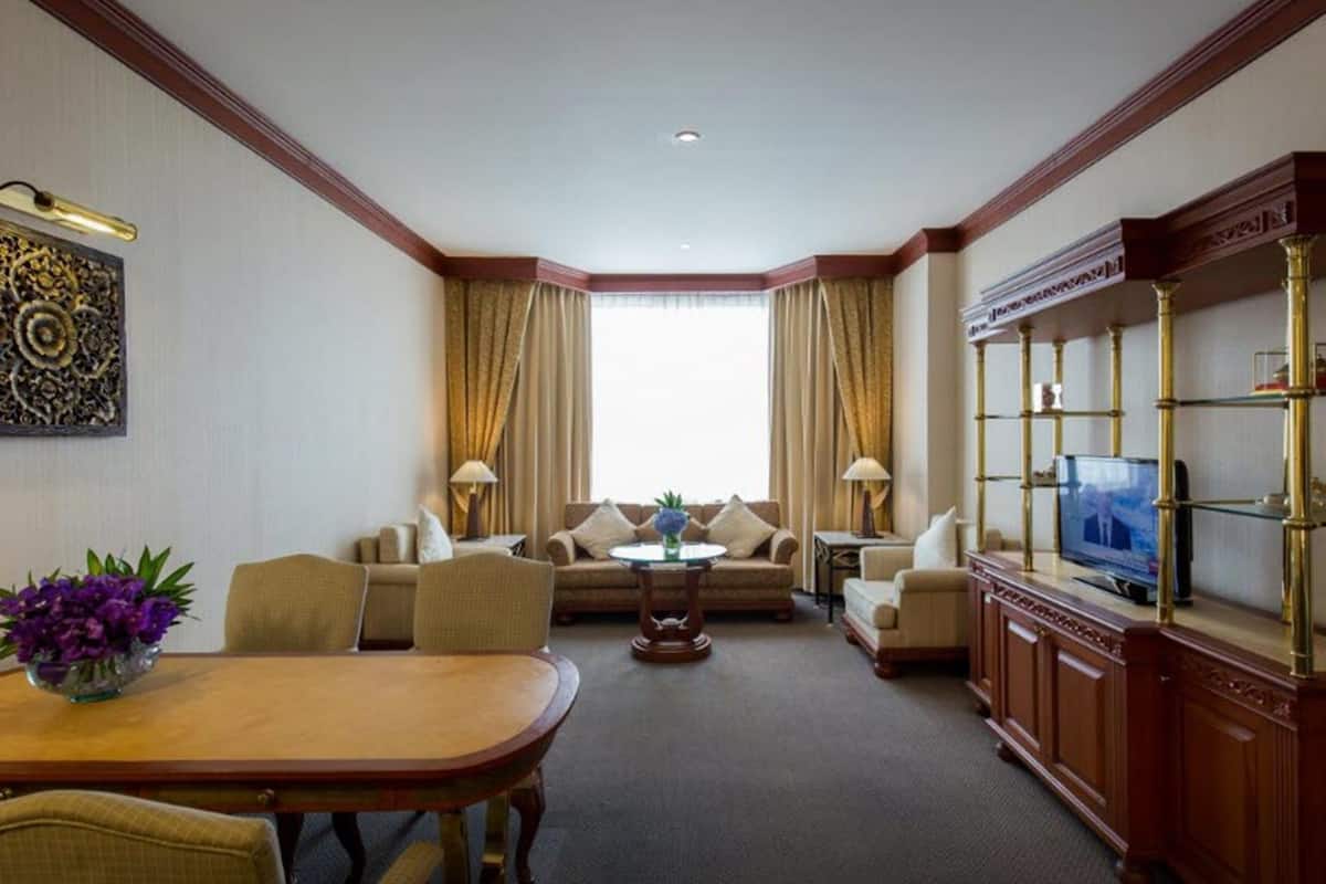 A hotel room with a dining area, a sofa set, a television on a wooden cabinet, and a large window with curtains.