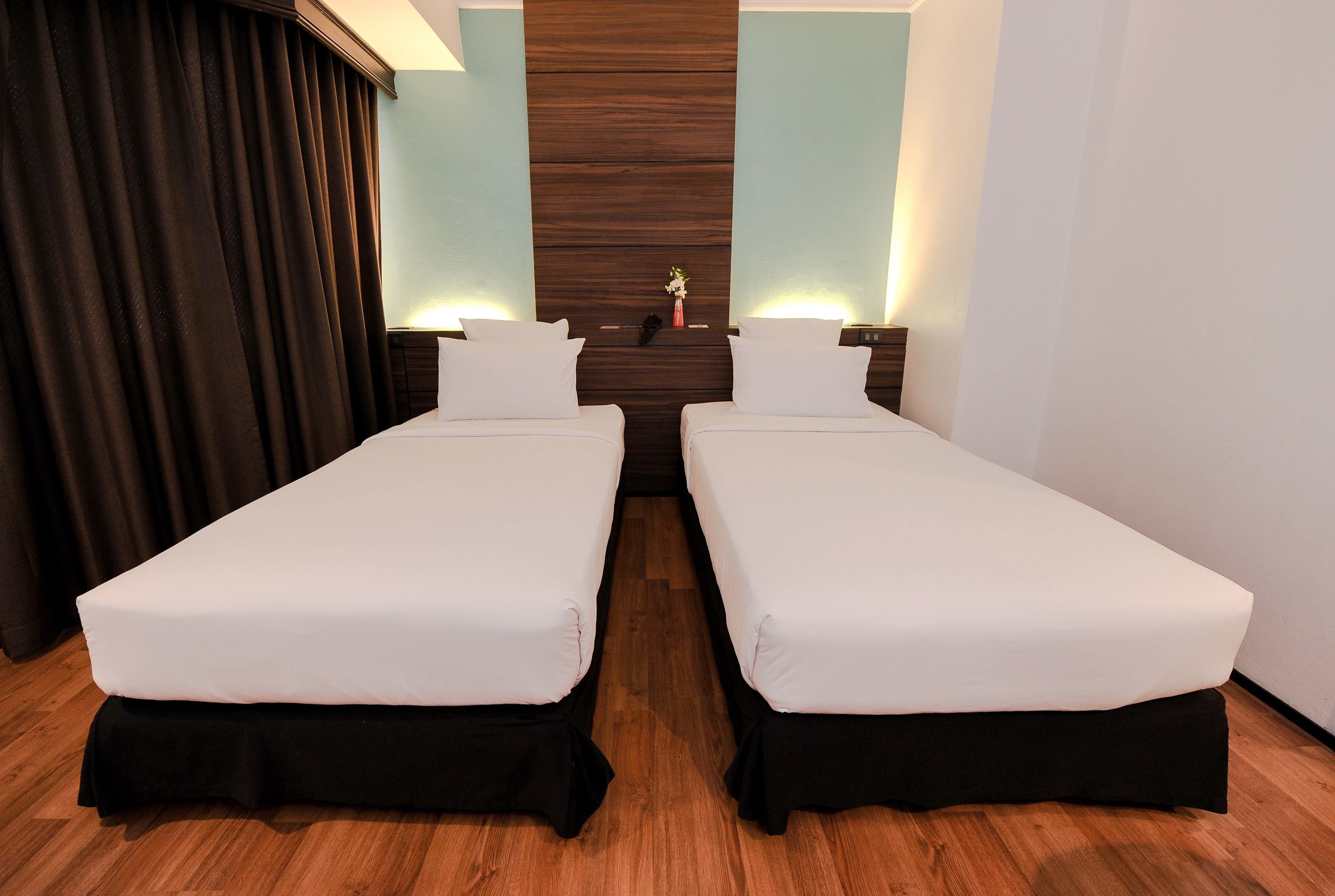 A hotel room with two single beds, wooden flooring, a dark curtain, a wooden accent wall, and bedside lamps.