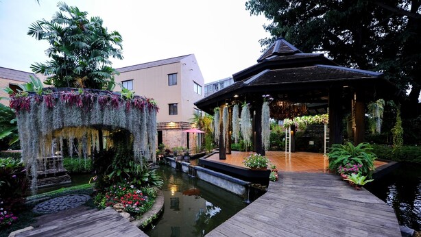 Une terrasse en bois avec un petit étang, un belvédère et une fontaine en pierre, le tout entouré d’une végétation luxuriante et de fleurs.