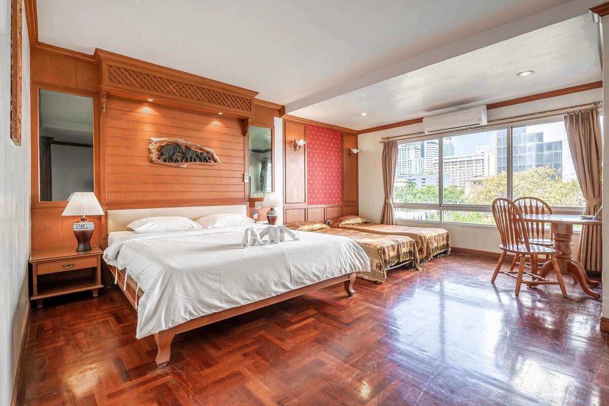 Une chambre spacieuse avec un grand lit, une tête de lit en bois, une table de chevet, une table avec des chaises et une fenêtre donnant sur des immeubles.