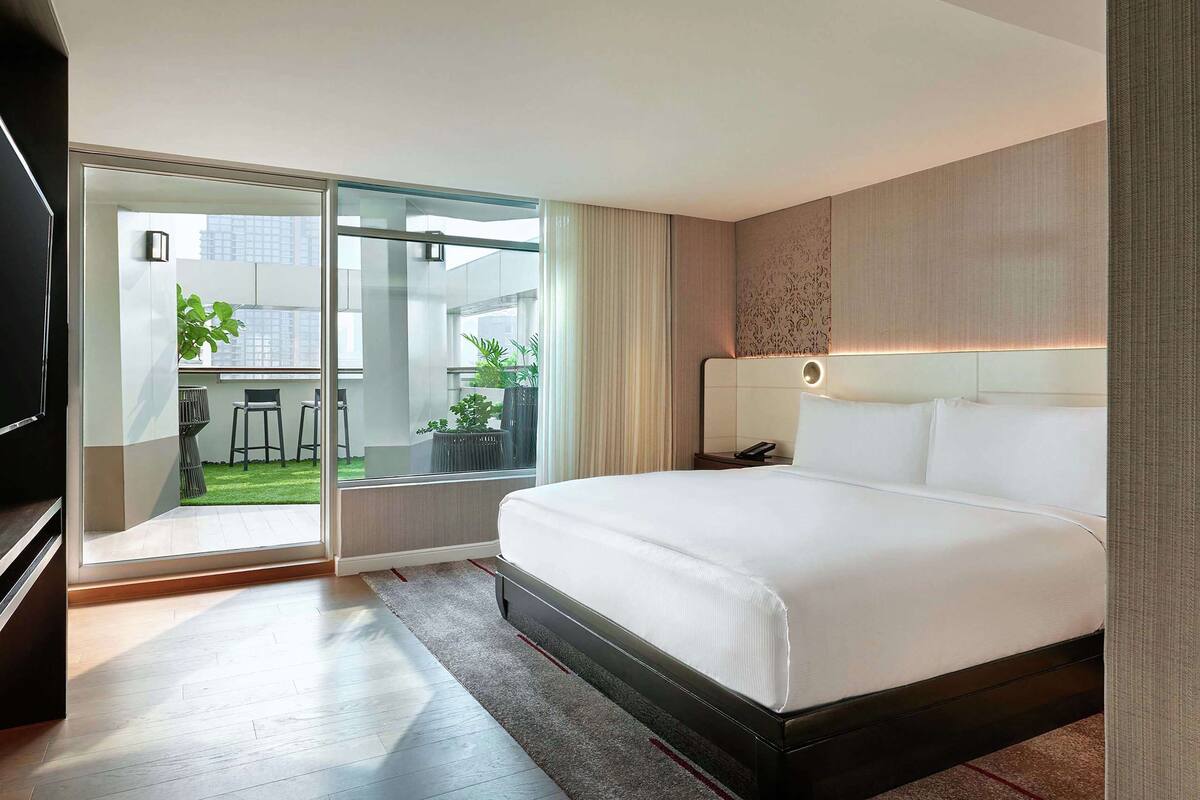 Une chambre d’hôtel moderne dotée d’un grand lit, d’une porte-fenêtre coulissante donnant sur un balcon avec vue sur la ville, et d’un petit coin salon avec une table et des chaises.