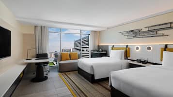 A hotel room with two beds, a desk, a TV, and a view of a cityscape.