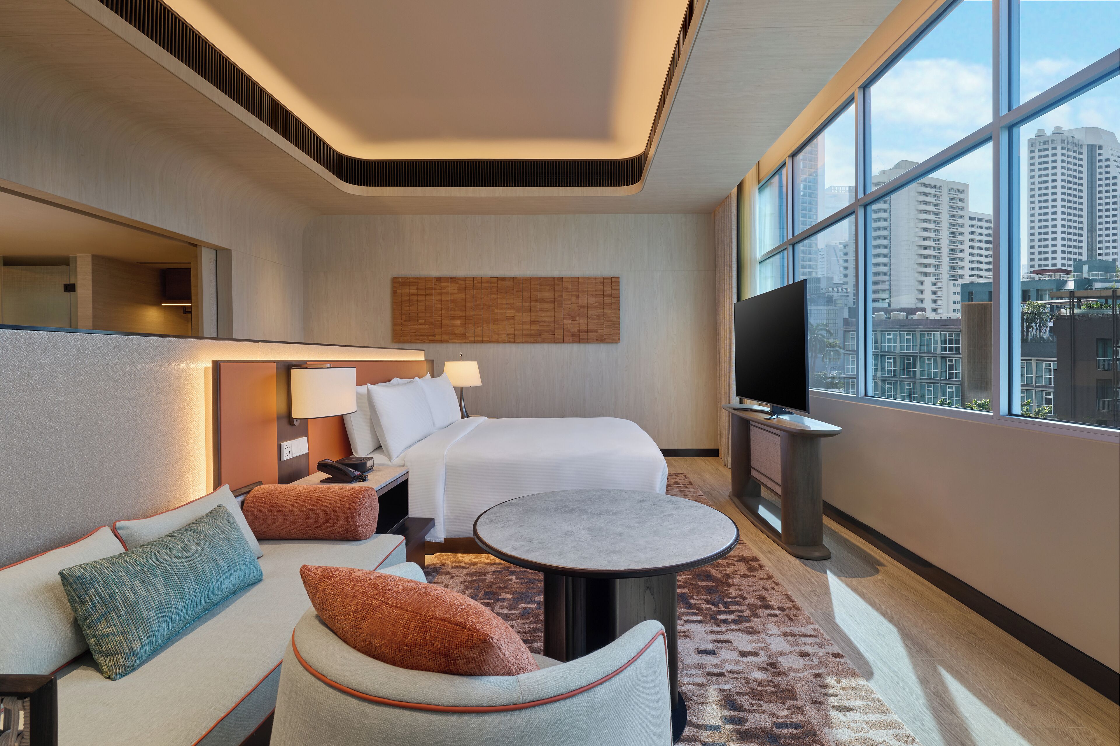 A modern hotel room with a bed, sofa, and TV, featuring a city view through large windows.