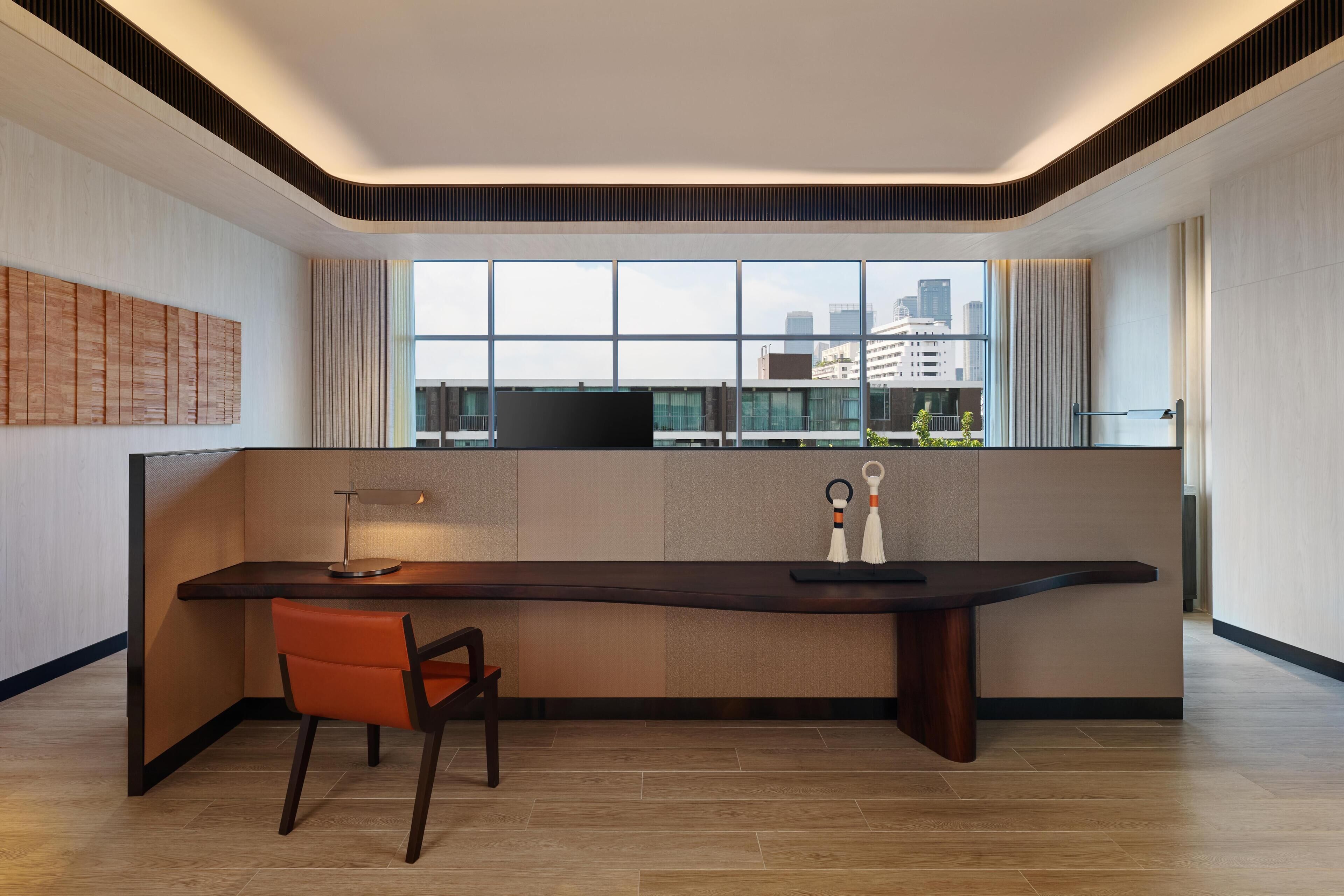A modern room with a long desk, a chair, a window with a city view, and a wall-mounted TV.