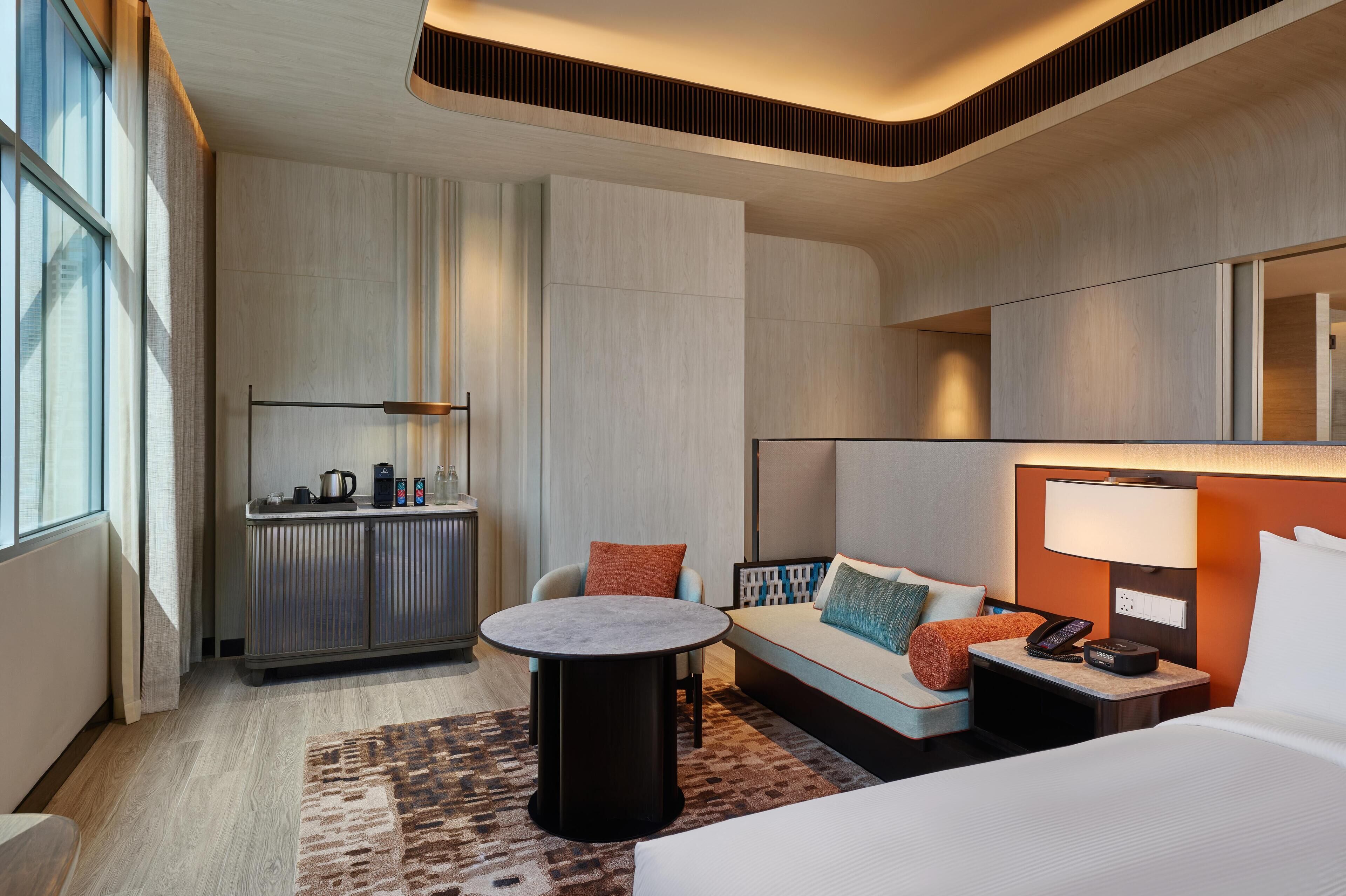A modern hotel room with a bed, a sofa, a small round table, a minibar, and a large window.