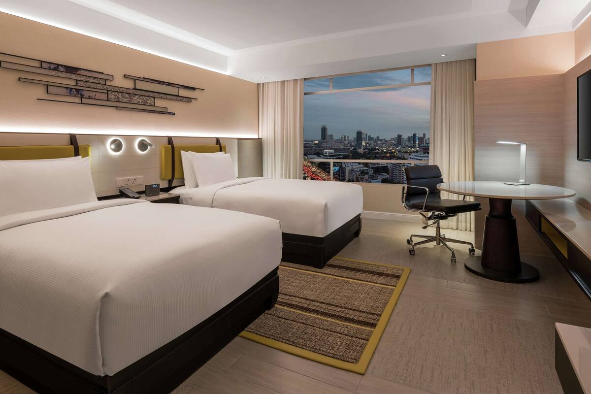 Une chambre d’hôtel avec deux lits, un bureau avec une chaise, une grande fenêtre donnant sur la ville et un téléviseur fixé au mur.
