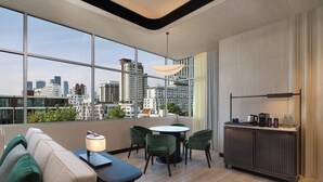 Een moderne hotelkamer met een groot raam, een ronde tafel, groene stoelen, een bank en een minibar.