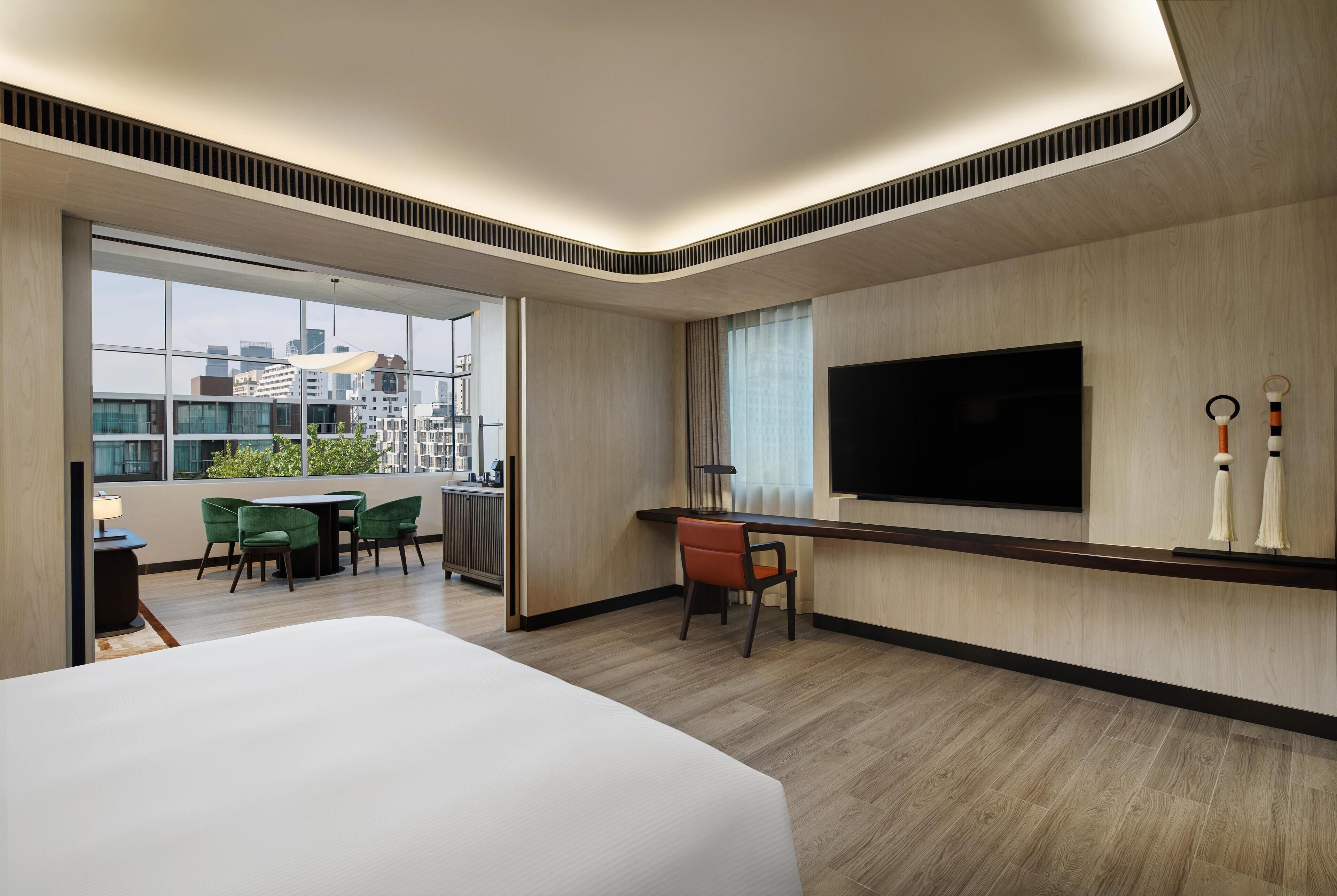 A modern hotel room with a large bed, a dining area with green chairs, a TV mounted on the wall, and a city view through the window.