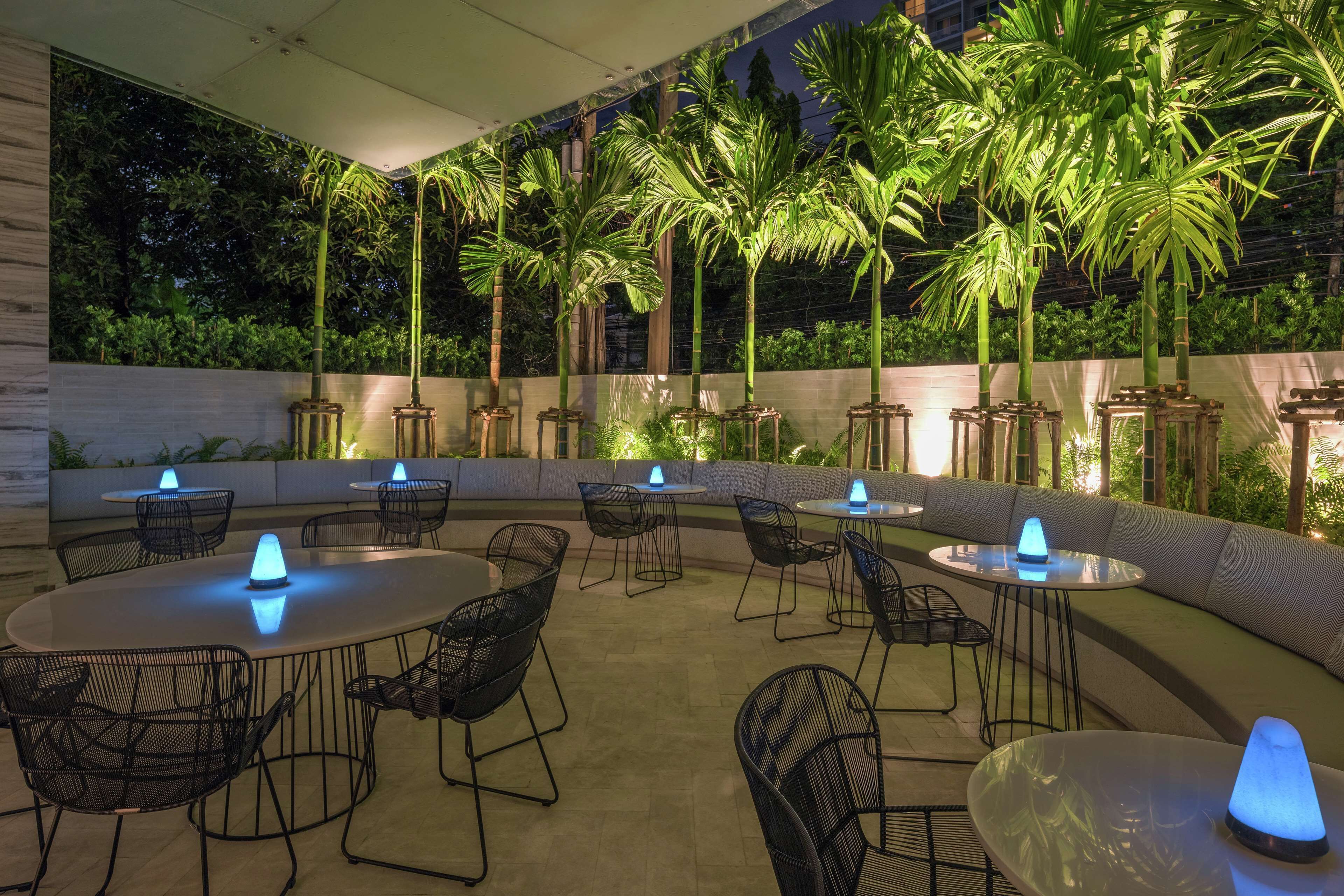 An outdoor seating area with round tables and chairs, surrounded by palm trees and a low wall.