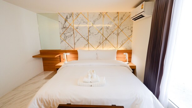 Une chambre d’hôtel avec un mur décoré d’un motif imitant le marbre, une tête de lit en bois, un lit recouvert de draps blancs et une table de chevet avec une lampe.