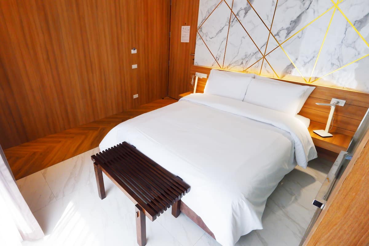 A hotel room with a wooden wardrobe, a bed with white bedding, a marble-patterned wall, and a small wooden bench.