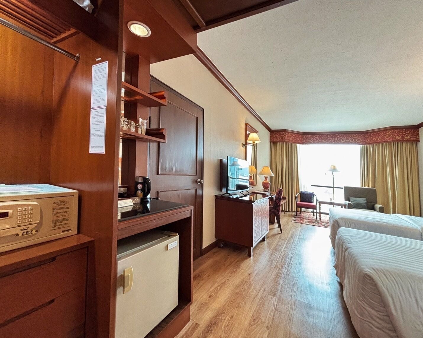 A hotel room with a wooden desk, a minibar, a television, a bed, and a view of the outdoors.