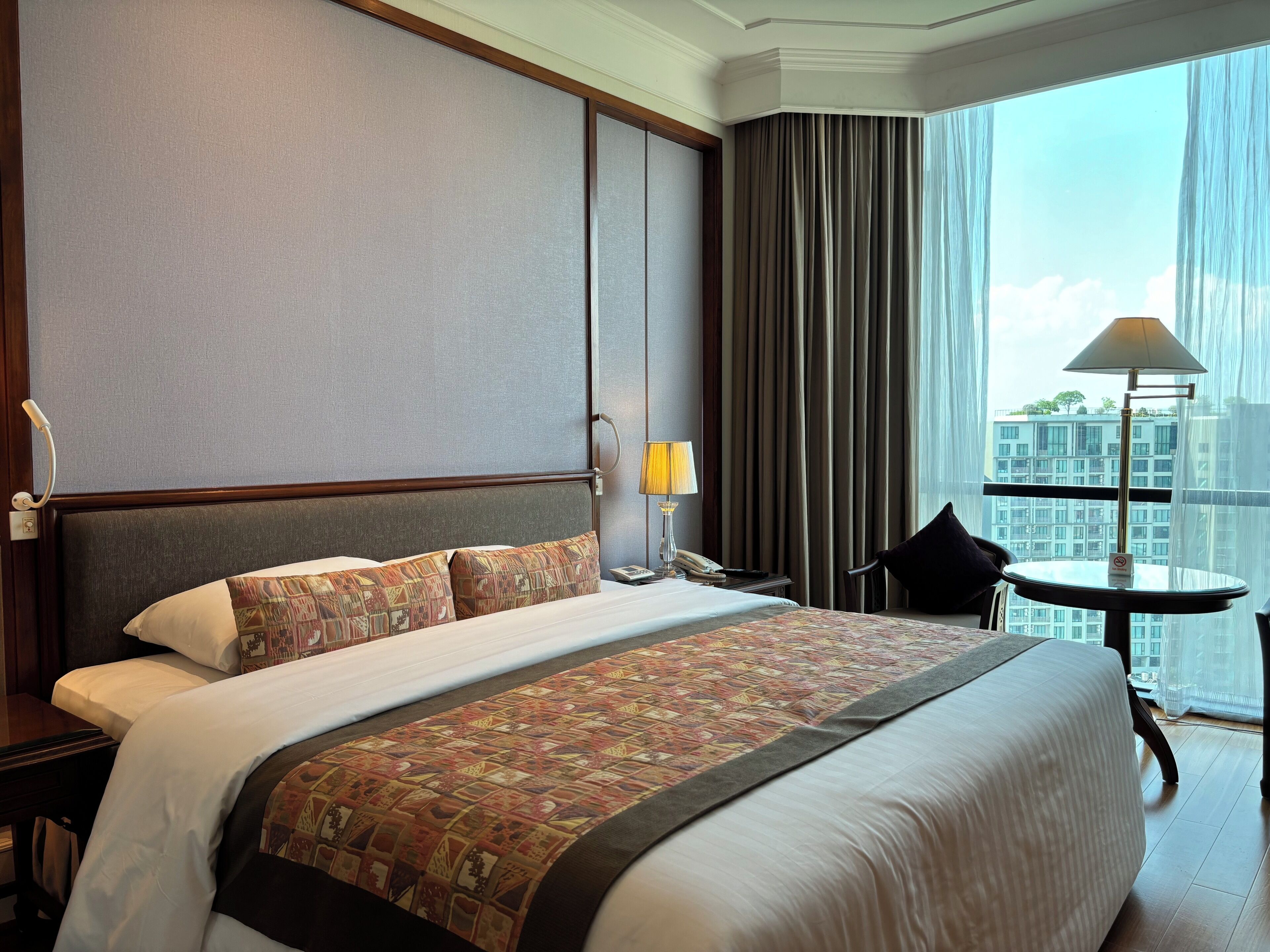 A hotel room with a large bed, a nightstand with a lamp, a chair, and a view of a cityscape through sheer curtains.