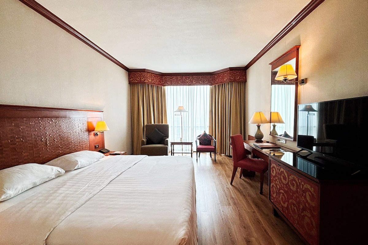 Quarto de hotel com uma cama grande, uma escrivaninha com televisão, uma cadeira e uma janela com cortinas.