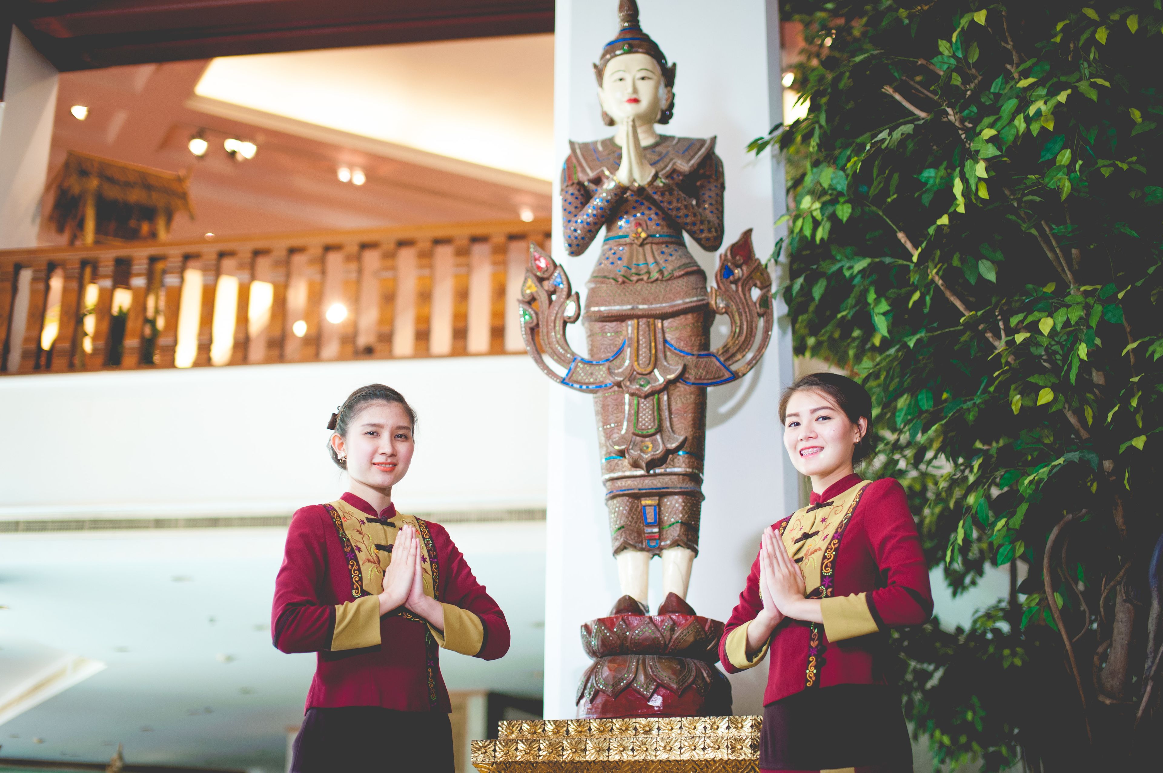 Two individuals in traditional attire standing beside a large, ornate statue in a well-lit indoor space with a wooden railing and greenery in the background.