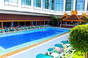 Zona de piscina con sillas de descanso y sombrillas, piscina, y edificio con ventanales grandes y una zona de estar cubierta.