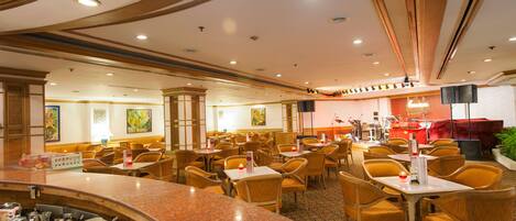 A hotel dining area with multiple tables and chairs, a bar with various food and drink items, and a stage with musical instruments.