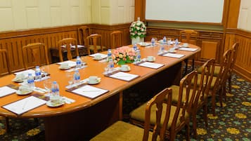 Un salón de conferencias con una mesa larga, sillas, botellas de agua, vasos y un arreglo floral.