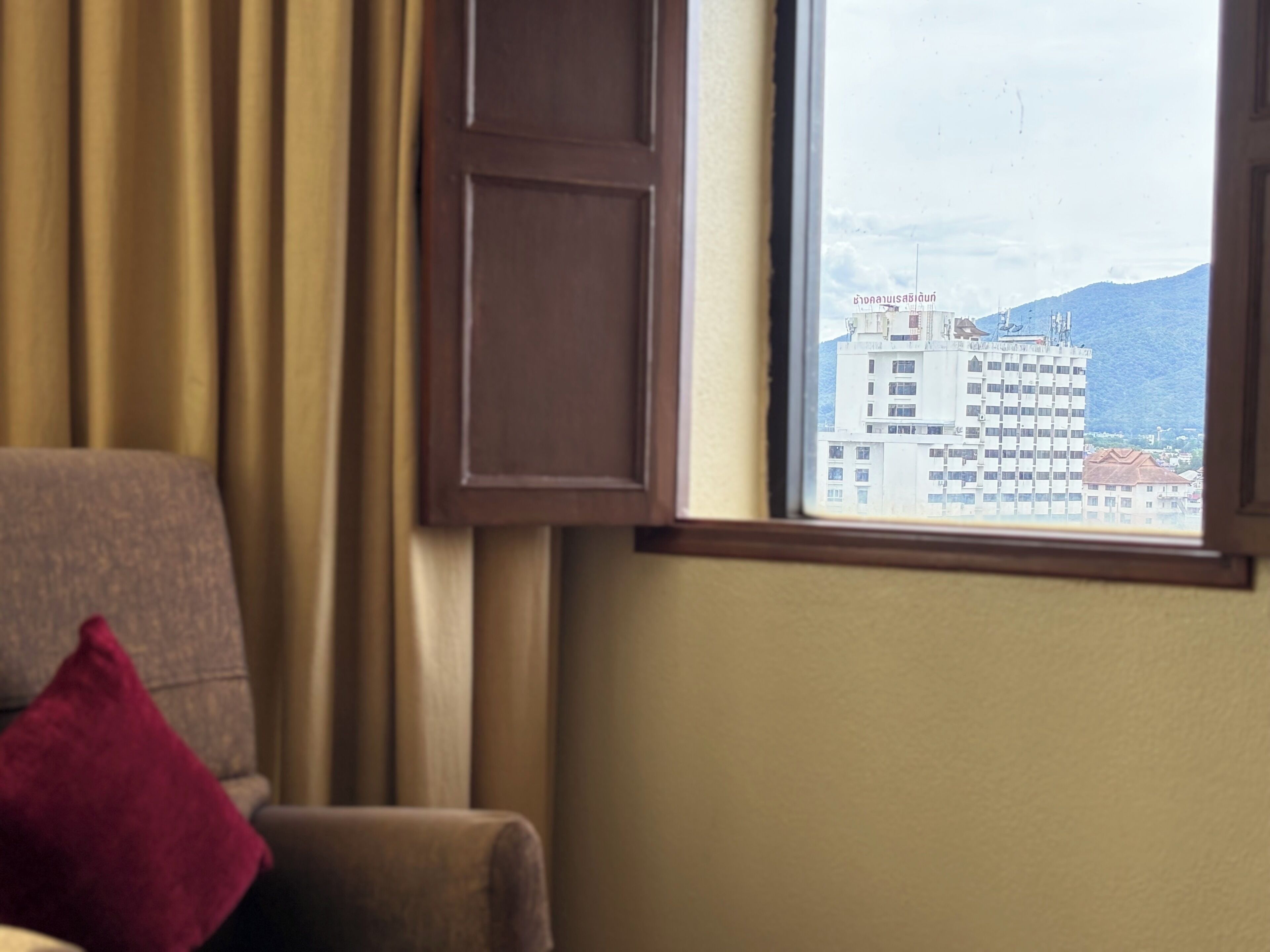 Una habitación con vista a un edificio a través de una ventana, una silla con un cojín rojo y una cortina.
