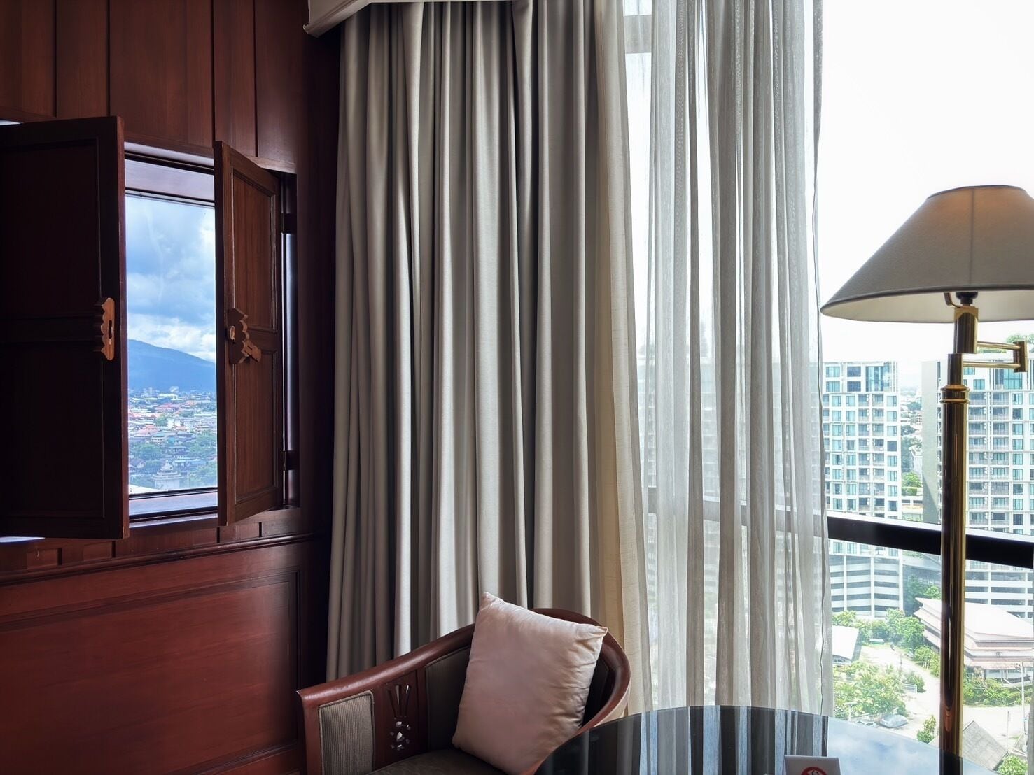 A room with a view of a cityscape, a wooden cabinet, a chair with a cushion, and a lamp.