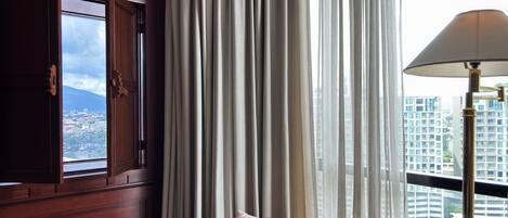 A room with a view of a cityscape, a wooden cabinet, a chair with a cushion, and a lamp.