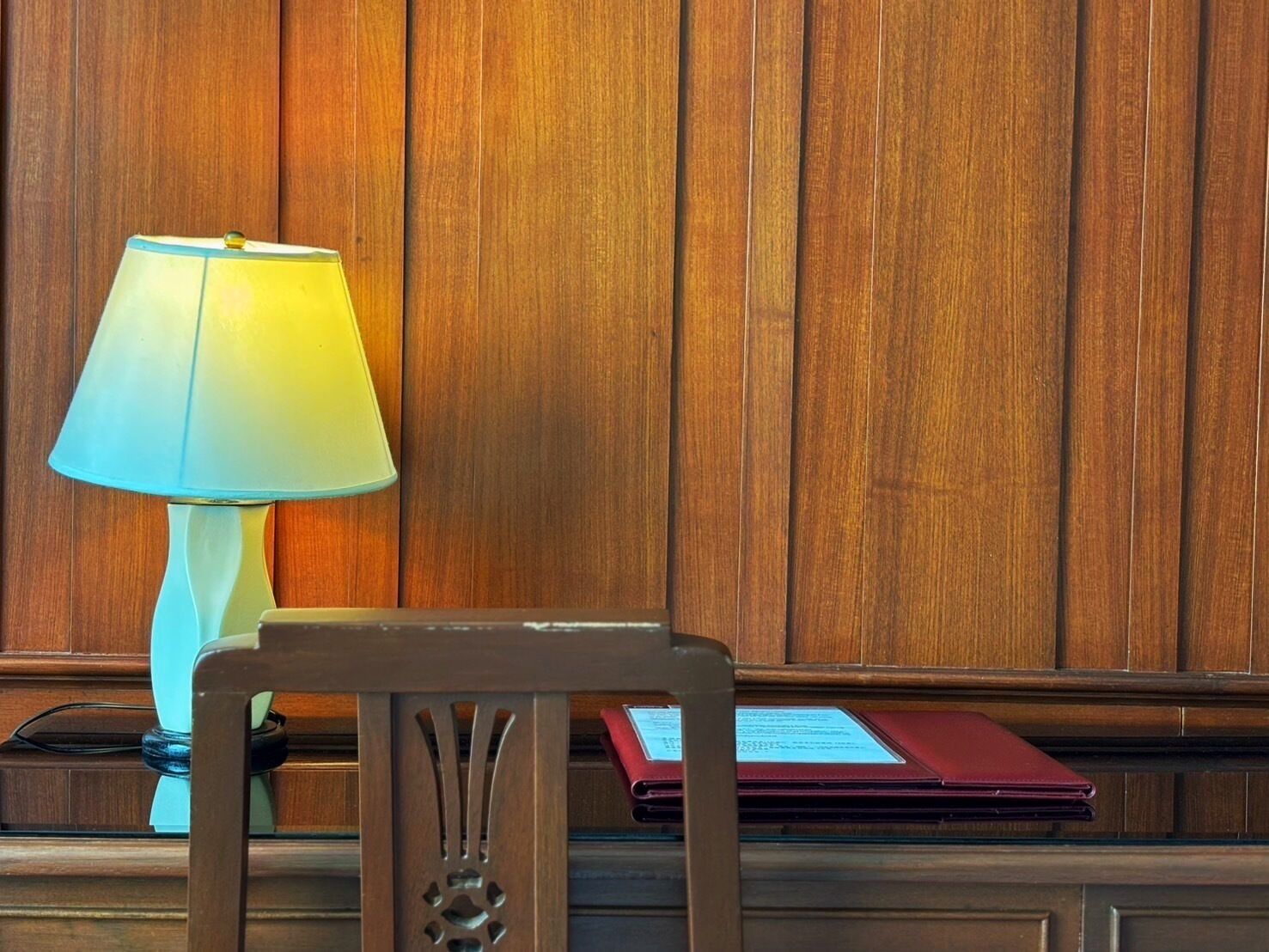 A wooden panel wall, a lamp, and a chair with a red folder on a desk.
