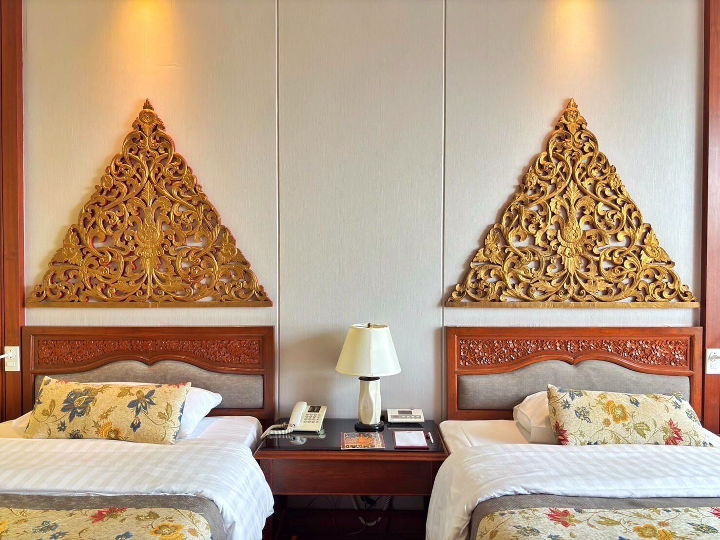 A hotel room with two beds, each with floral-patterned bedding and headboards. There is a bedside table with a lamp, a phone, and a book.