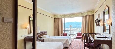 A hotel room with two beds, a desk, a chair, a large mirror, and a view of the city.
