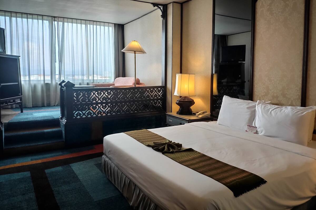 Ein Hotelzimmer mit einem großen Bett, einem Schreibtisch mit Lampe, einem Telefon und Blick ins Freie durch transparente Vorhänge.