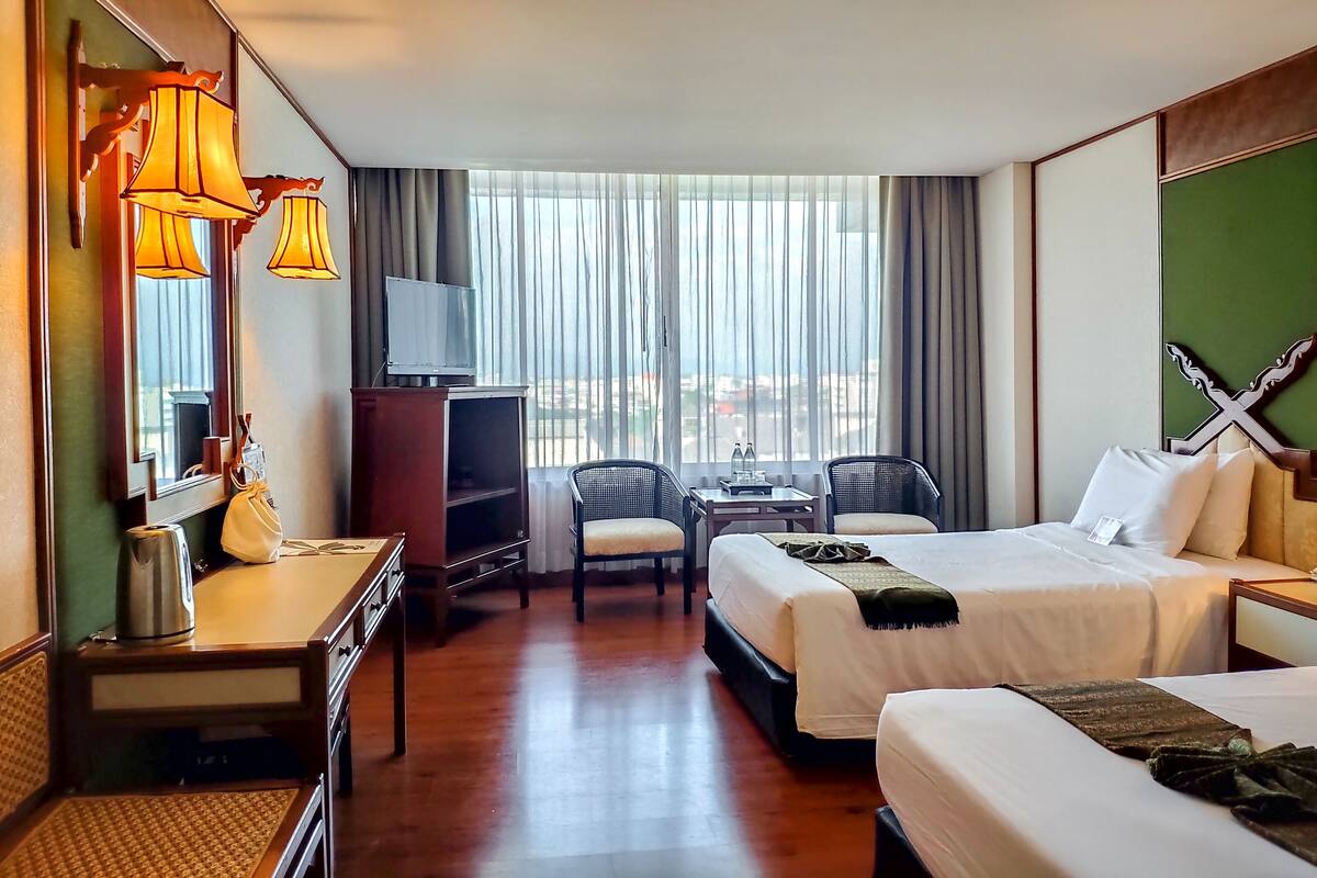 Ein Hotelzimmer mit zwei Betten, einem Schreibtisch, einem Fernseher und Blick auf die Stadt durch große Fenster.