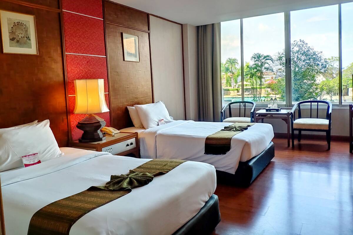 Ein Hotelzimmer mit zwei Betten, einem Schreibtisch, einem Stuhl und einem großen Fenster mit Blick ins Freie.