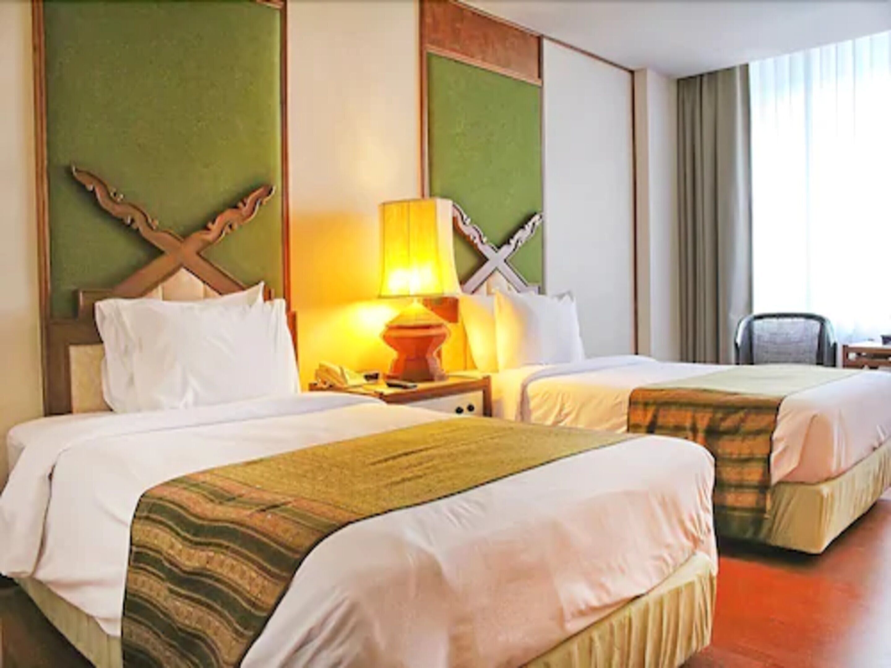 A hotel room with two beds, a nightstand with a lamp, and a chair.
