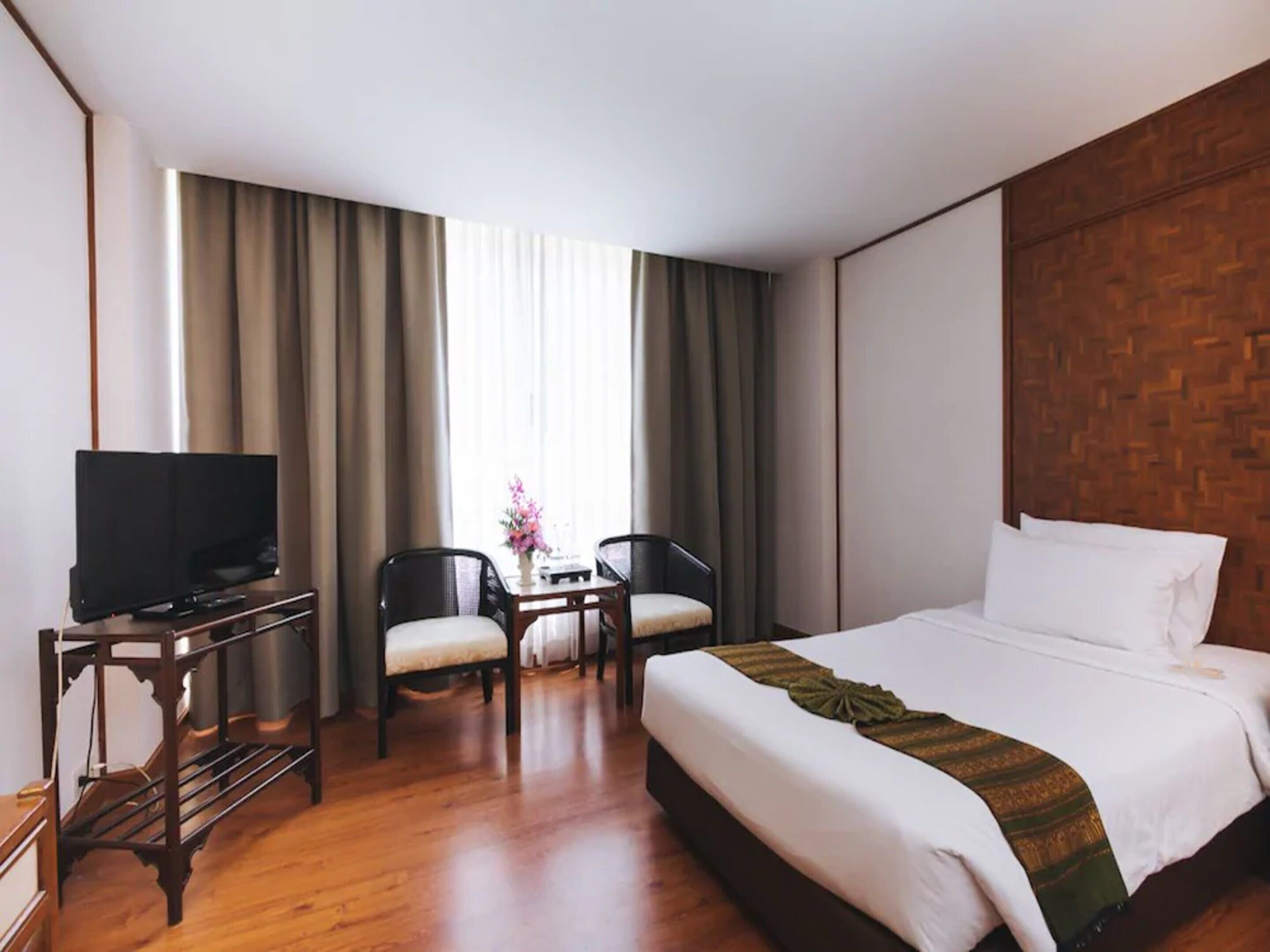 Una habitación de hotel con una cama grande, un televisor sobre un mueble de madera, dos sillas, una mesita con un jarrón y una ventana con cortinas.