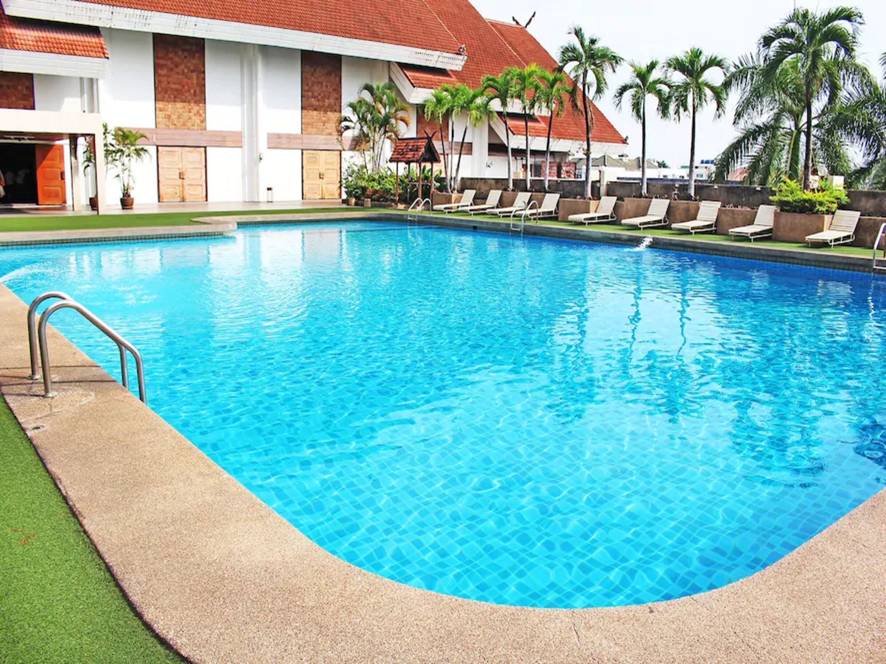 Ein Swimmingpool mit Liegestühlen und Palmen, umgeben von einem Gebäude mit rotem Dach.