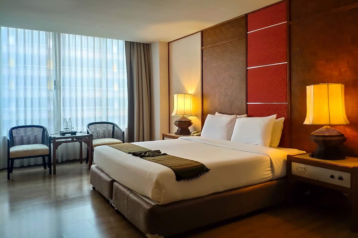 Ein Hotelzimmer mit einem großen Bett, zwei Nachttischen mit Lampen, einem kleinen Tisch mit Stühlen und einem großen Fenster mit Vorhängen.