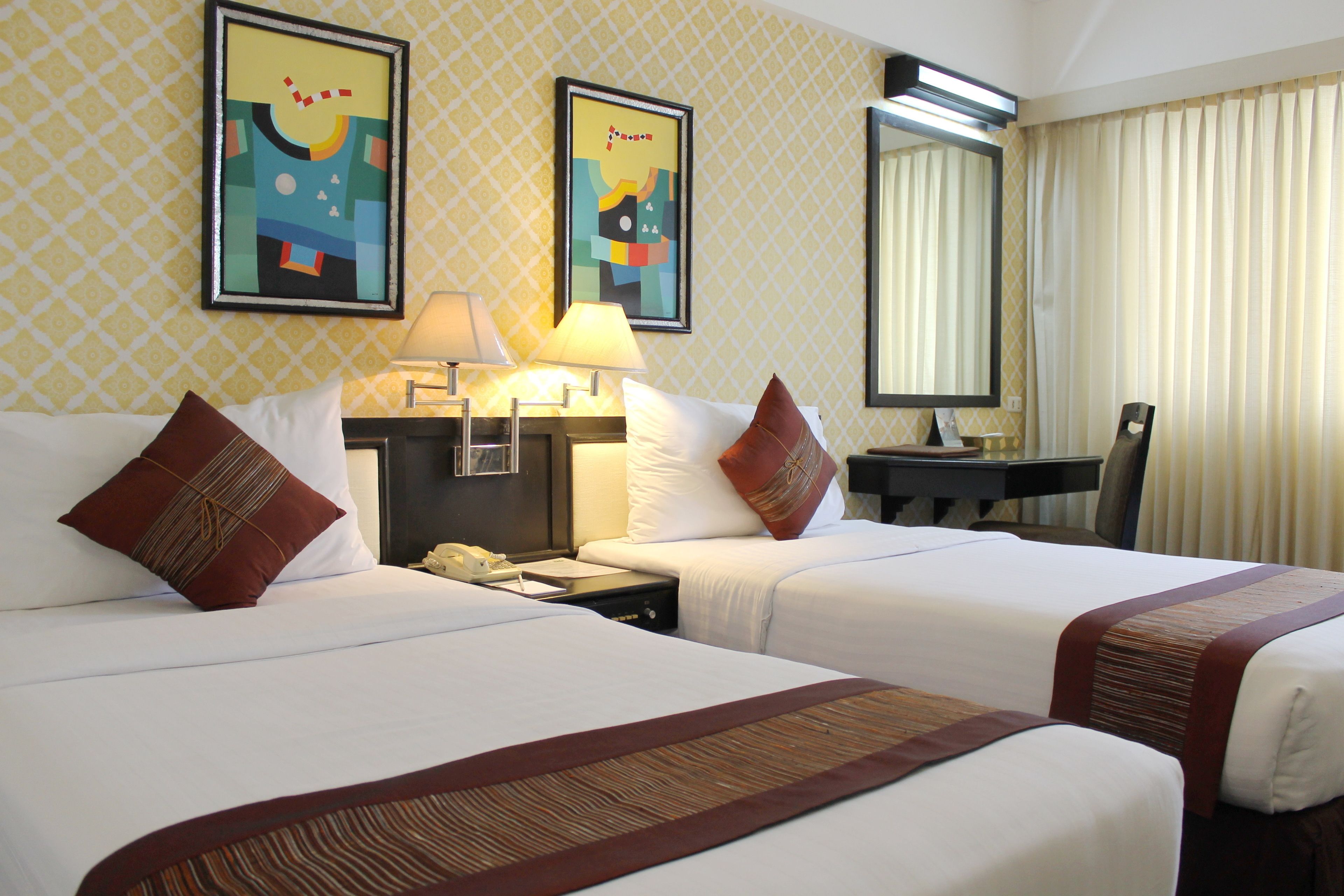 A hotel room with two beds, a desk with a lamp, a telephone, and two framed artworks on the wall.
