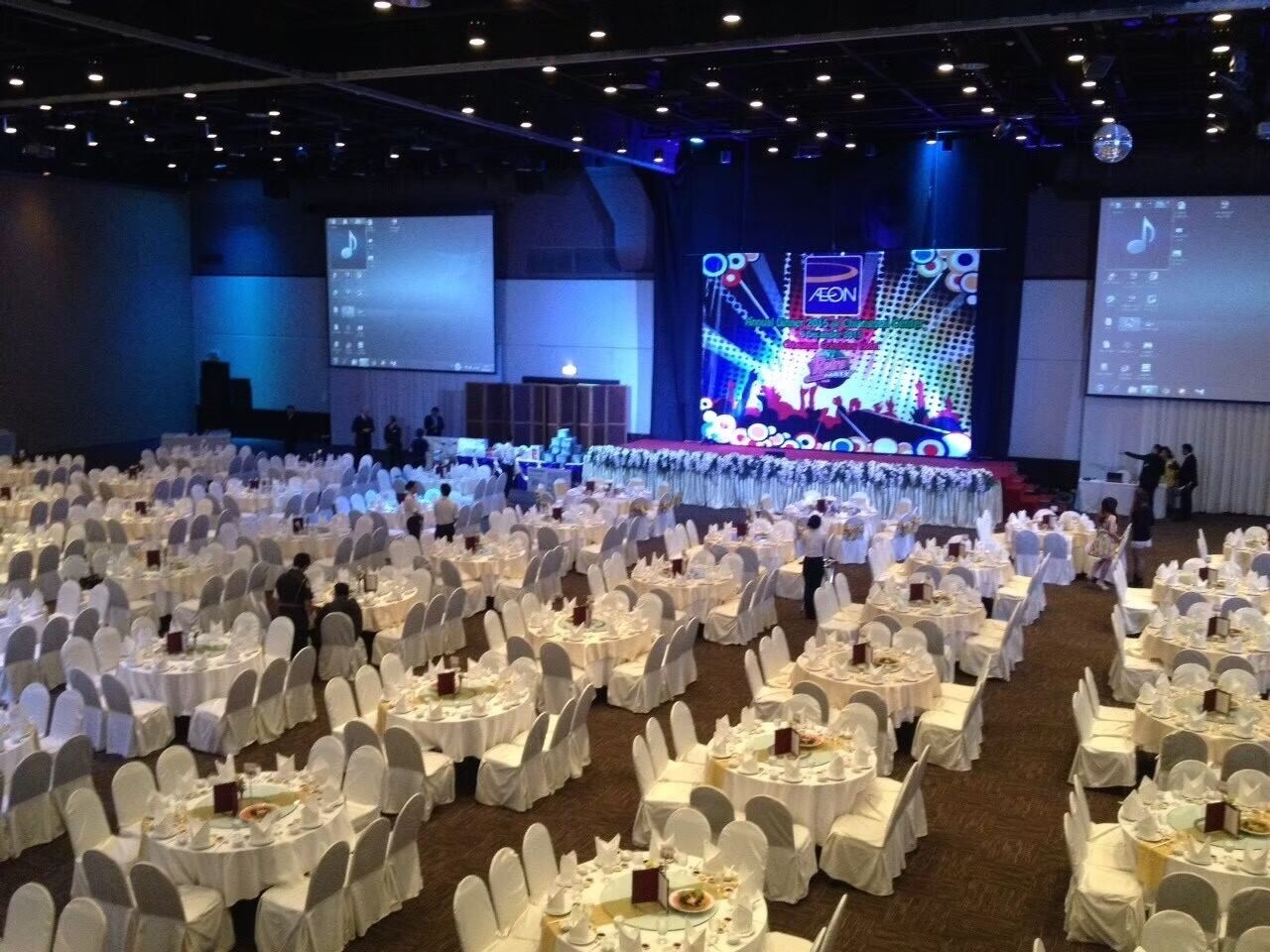 Uma sala de banquetes com mesas redondas cobertas por toalhas brancas e cadeiras, um palco com uma tela grande exibindo uma apresentação musical, e um chão coberto de carpete.