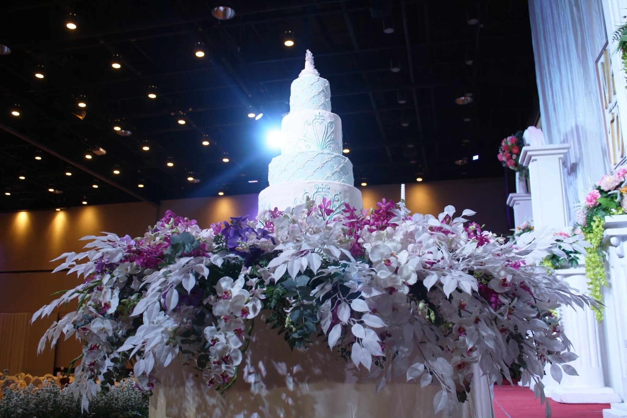A multi-tiered white cake with intricate icing designs, surrounded by a floral arrangement of white and purple flowers.