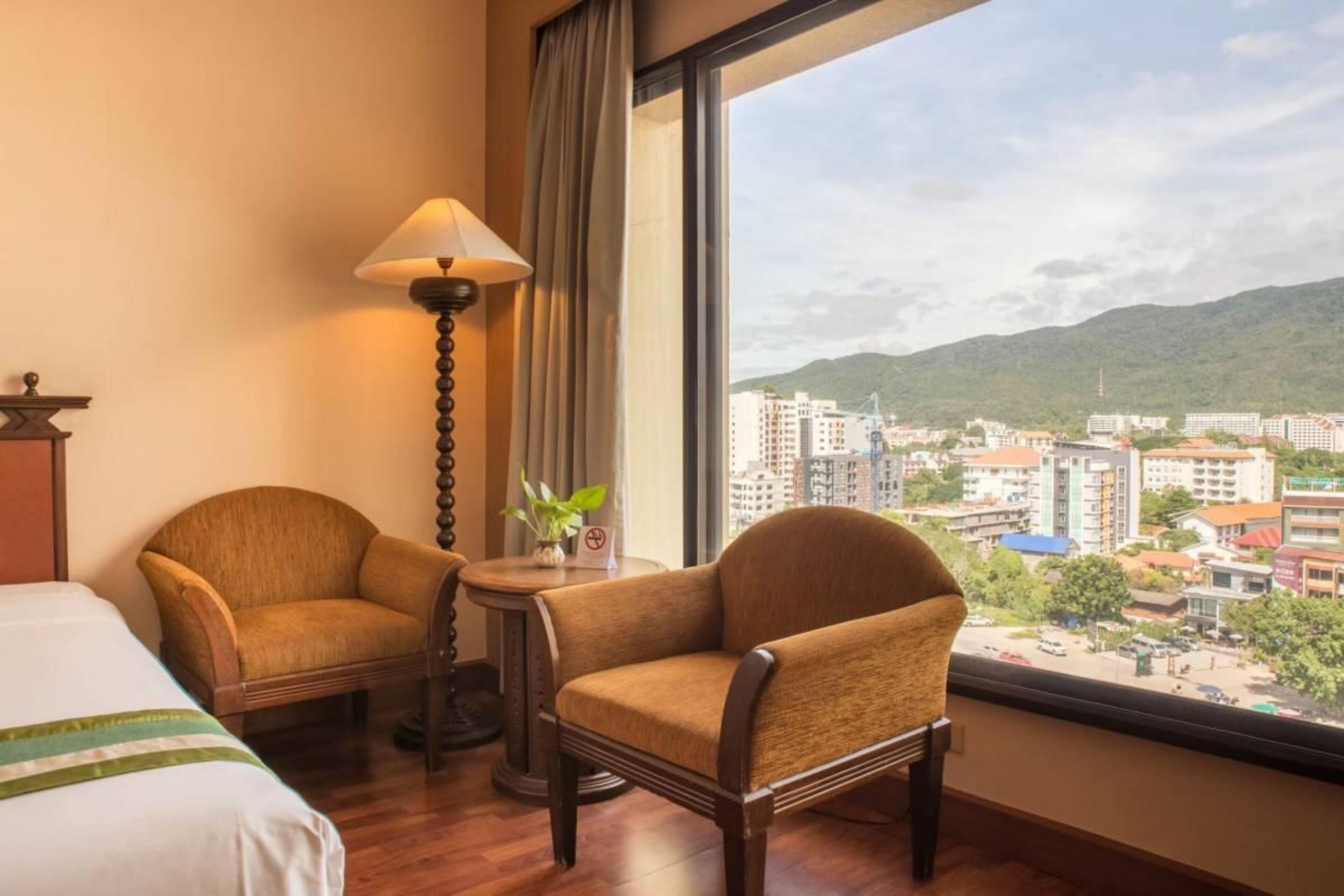 Habitación de hotel con una cama, dos sillones, una lámpara y vistas a un paisaje urbano a través de amplios ventanales.