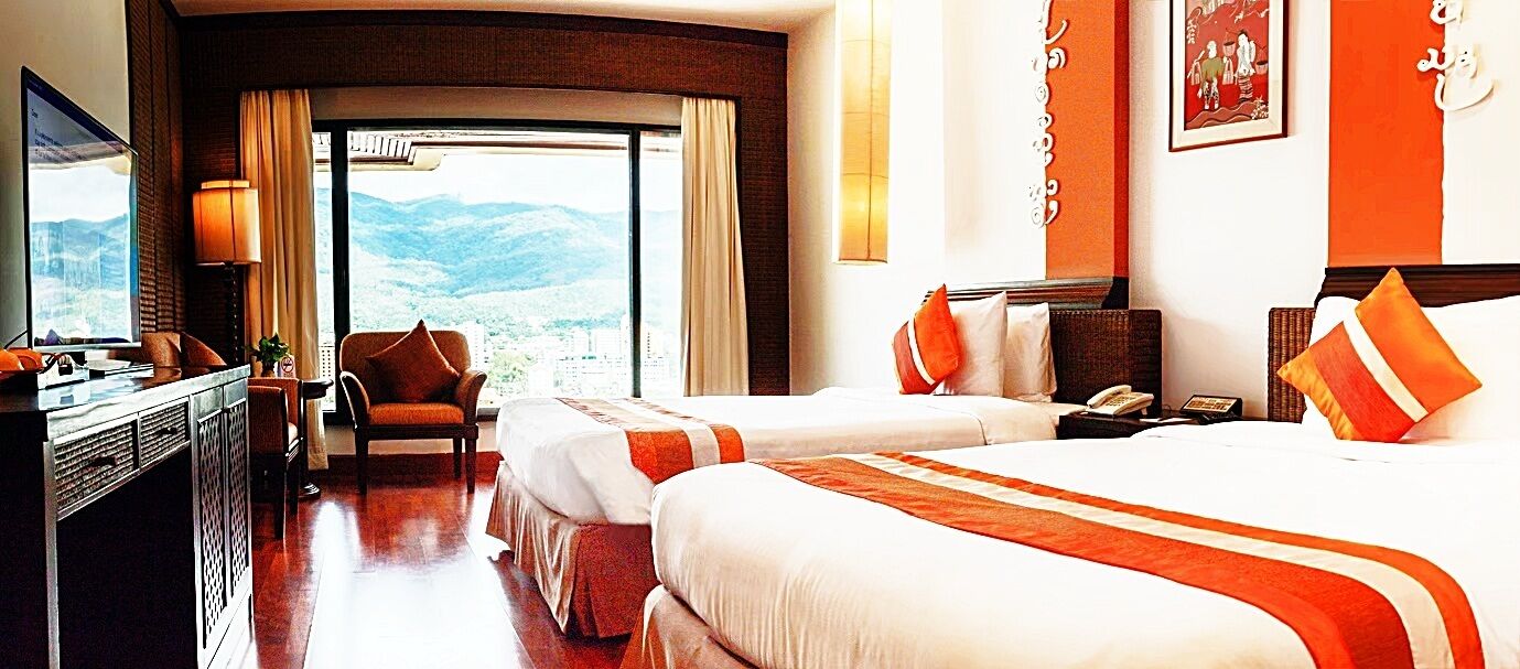 A hotel room with two beds, a desk, a chair, and a large window offering a mountain view.