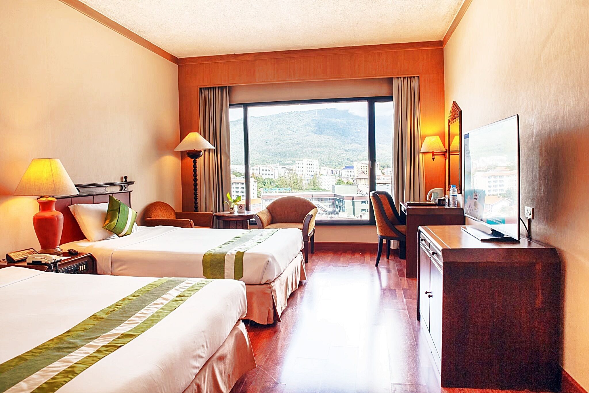 A hotel room with two beds, a desk, a chair, a TV, and a large window with a city view.