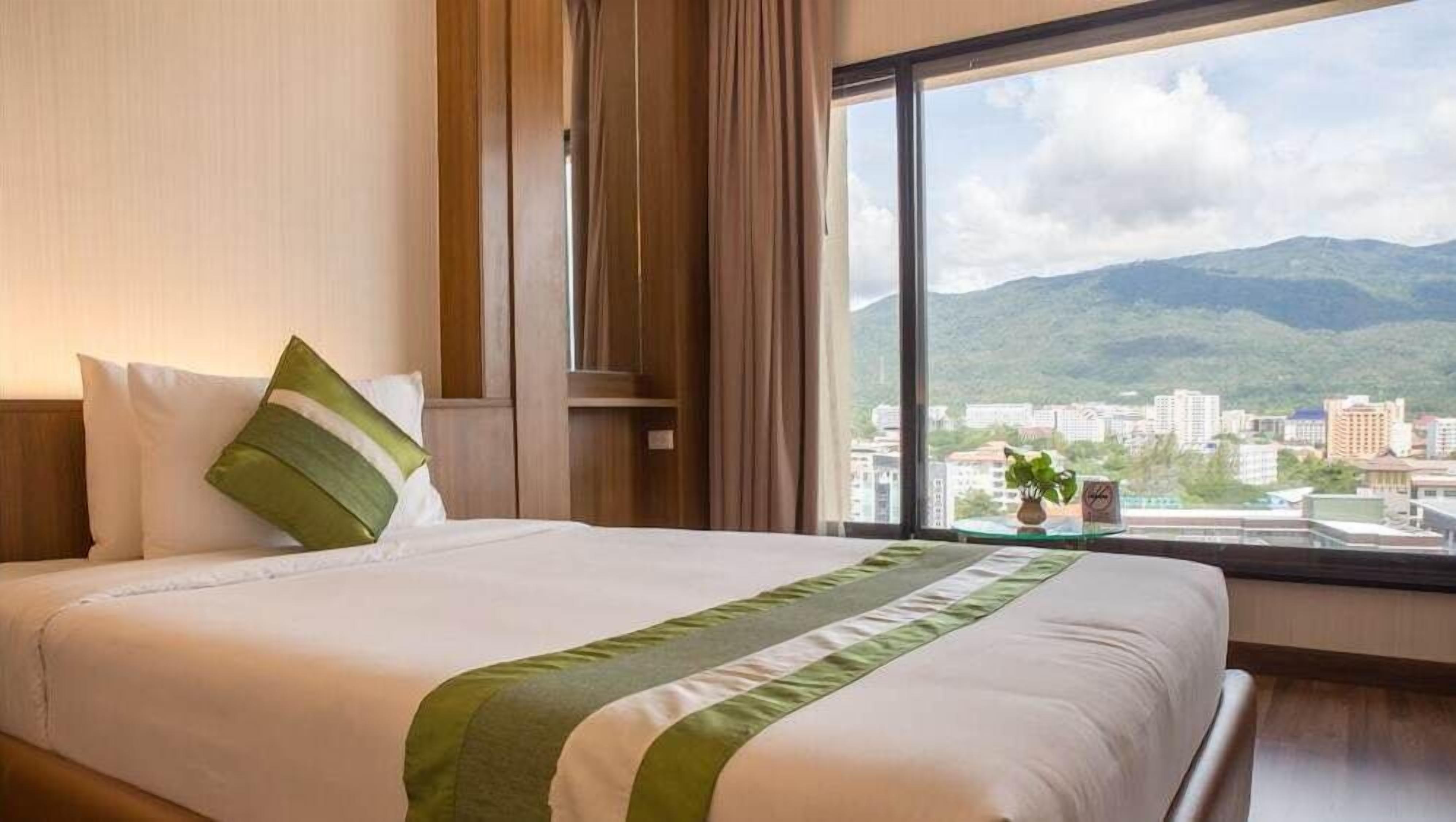 A hotel room with a large bed, a view of a cityscape and mountains, and a window with curtains.