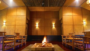 Una habitación con una chimenea central rodeada de sillas de madera, un muro con diseño y luces fijadas a la pared.