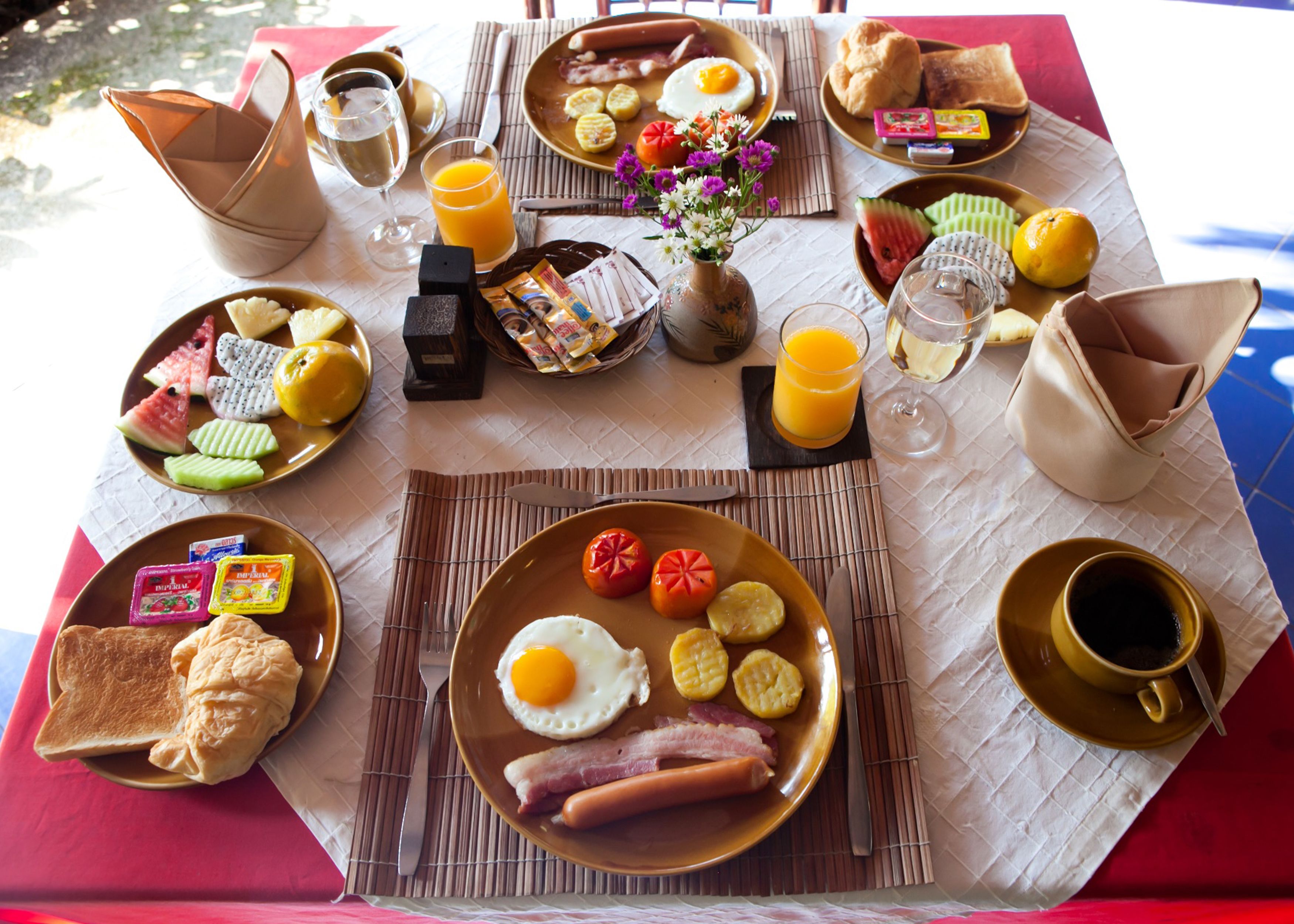 A breakfast table with plates of eggs, bacon, toast, and fruit. There are also glasses of orange juice and a vase with flowers.