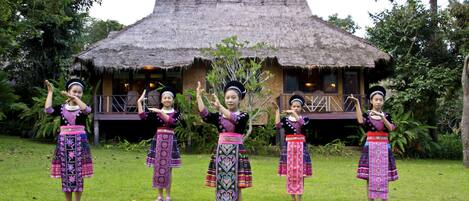 A group of people in traditional attire performing in front of a thatched-roof structure.
